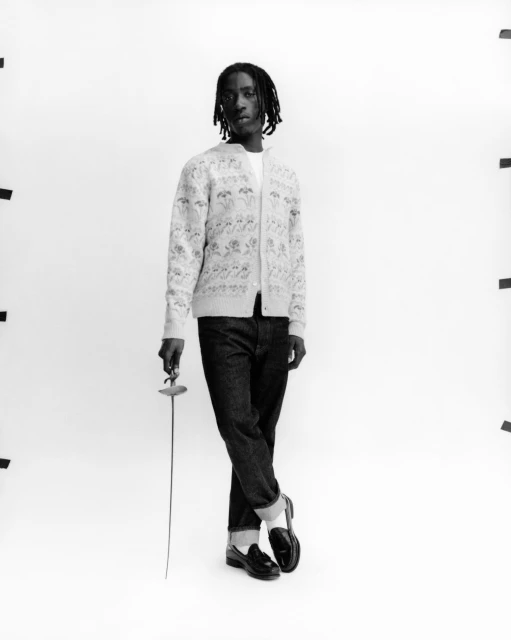 Black male fashion model posing in cardigan, jeans, and loafers, holding a fencing foil in a bright studio setting.