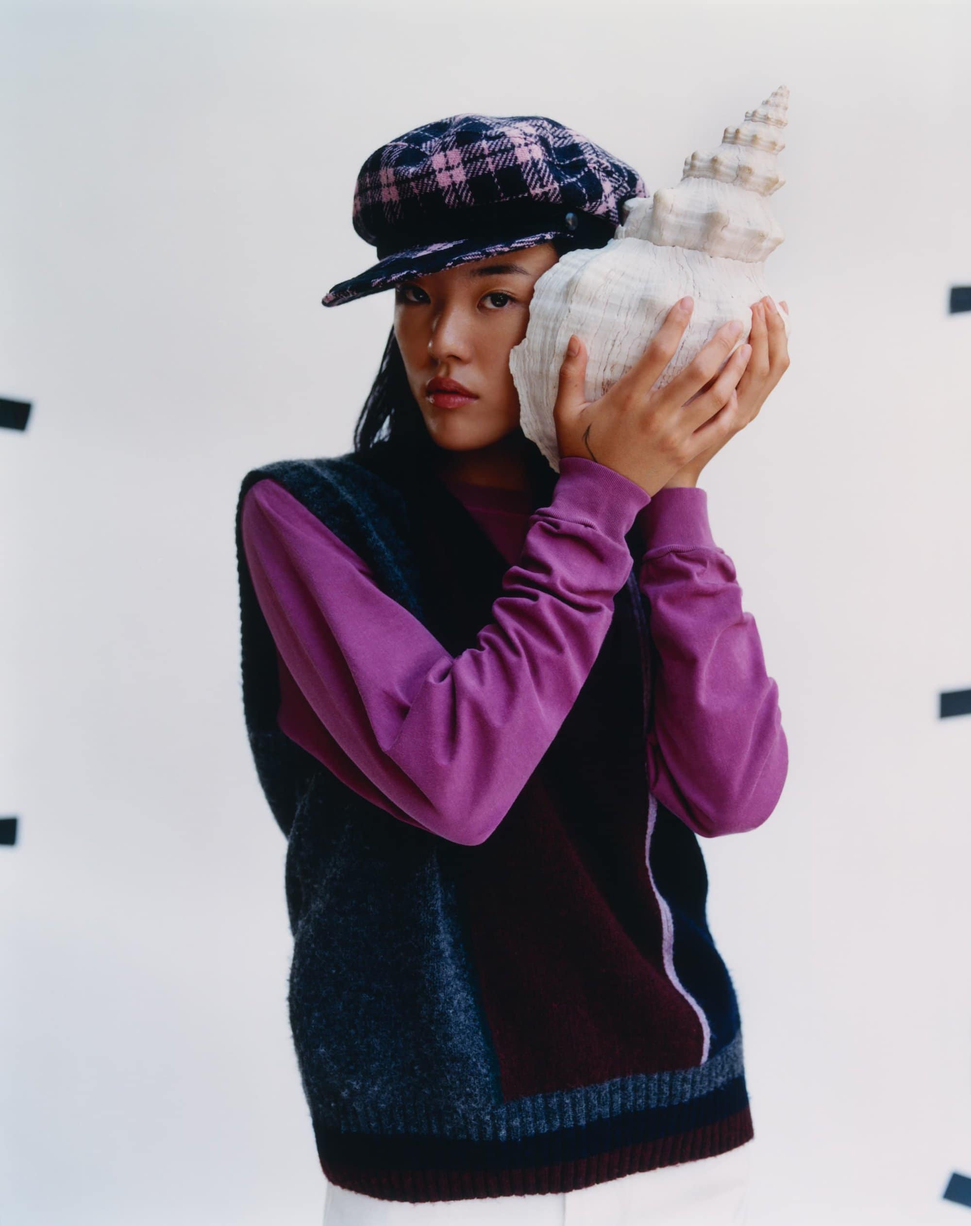 A young woman with a plaid newsboy cap holds a large seashell to her ear in a studio shot with a white background and diffused lighting.