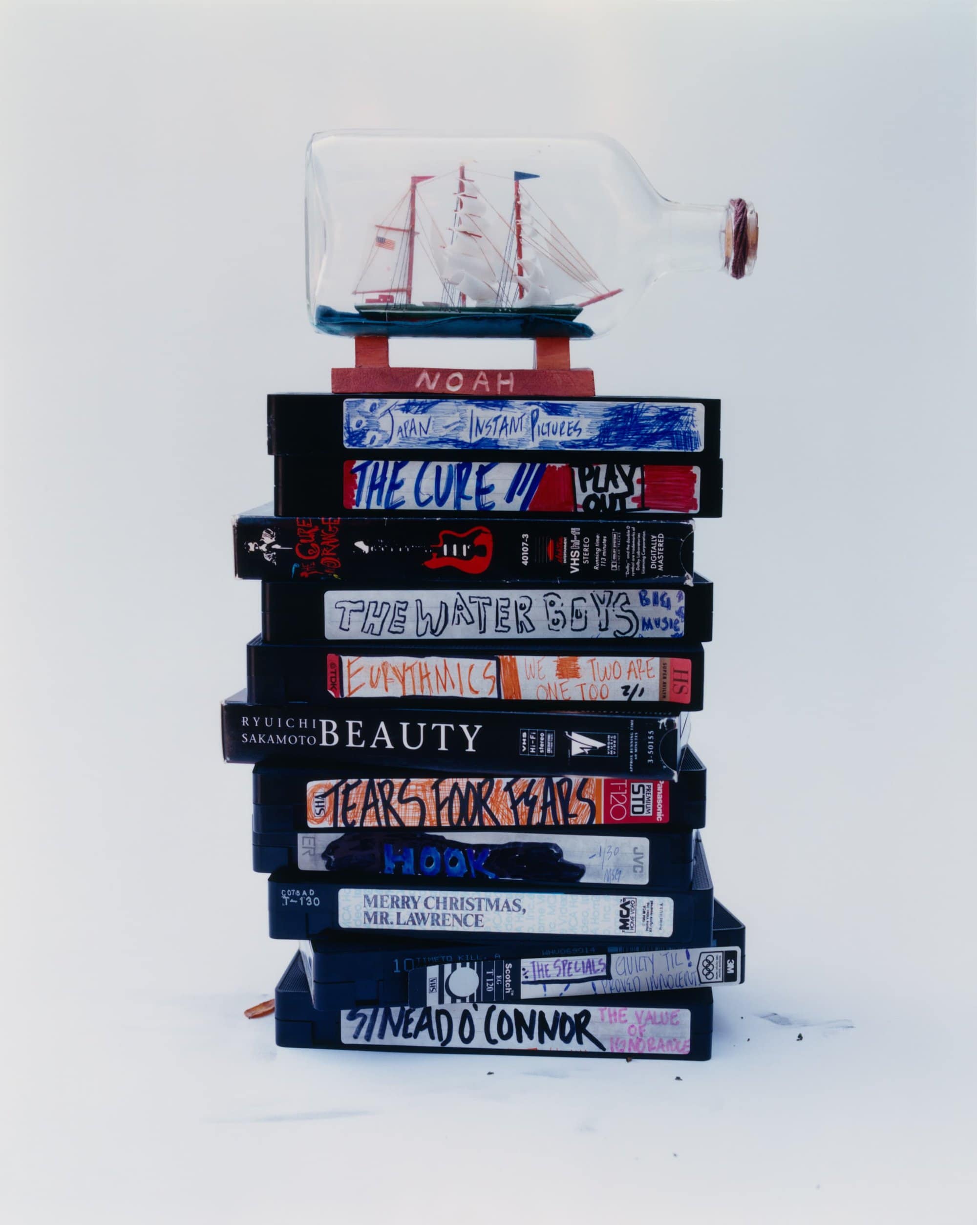 Still life of VHS tape stack, with a ship-in-a-bottle on top, against a white backdrop. Tapes have titles and drawn text on them.