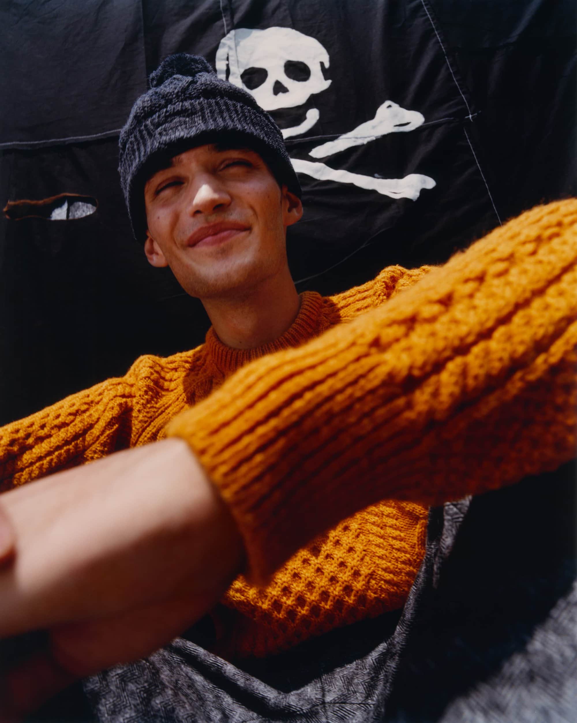 Portrait of a smiling young adult male wearing a knit hat and sweater, set against a black flag with a white skull and crossbones design.