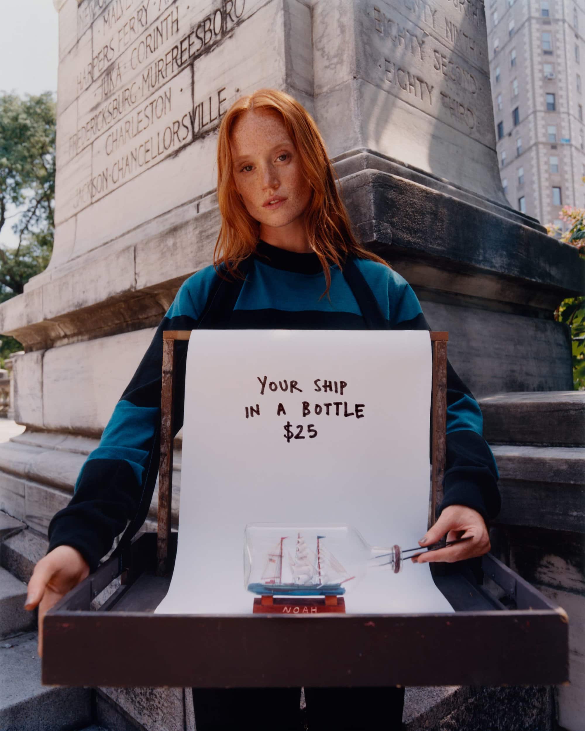 Adult female holding a ship in a bottle displayed on a tray with handwritten sign, set in an outdoor, urban environment.