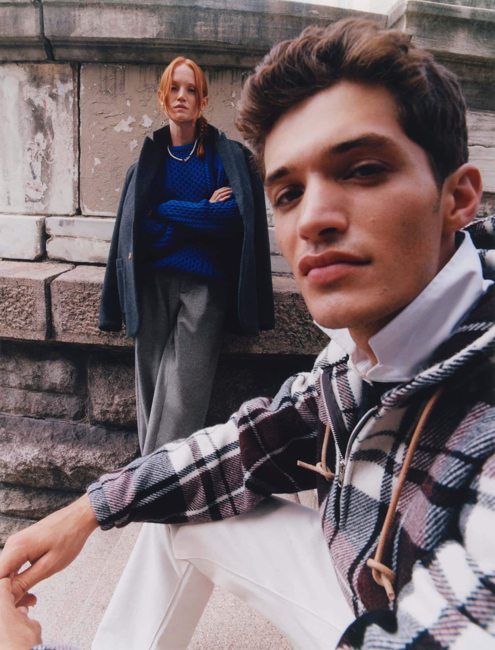A stylish outdoor fashion shoot featuring a young man in a plaid jacket in the foreground and a woman in a blue sweater behind him.
