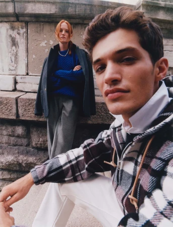 A stylish outdoor fashion shoot featuring a young man in a plaid jacket in the foreground and a woman in a blue sweater behind him.