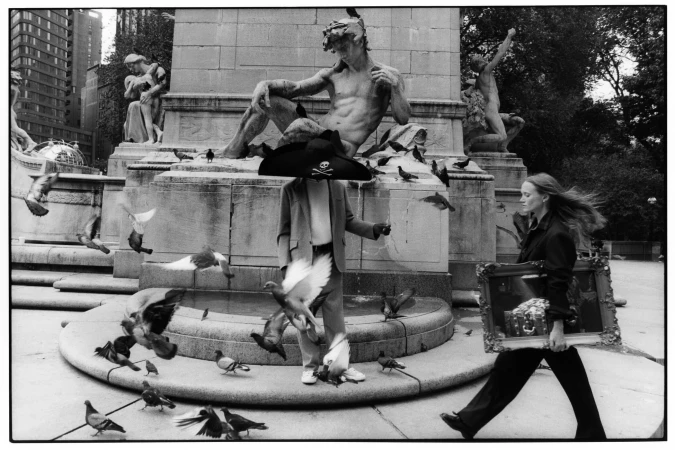 Black and white photograph of a public space featuring a woman carrying a framed artwork and a statue adorned with a pirate hat.