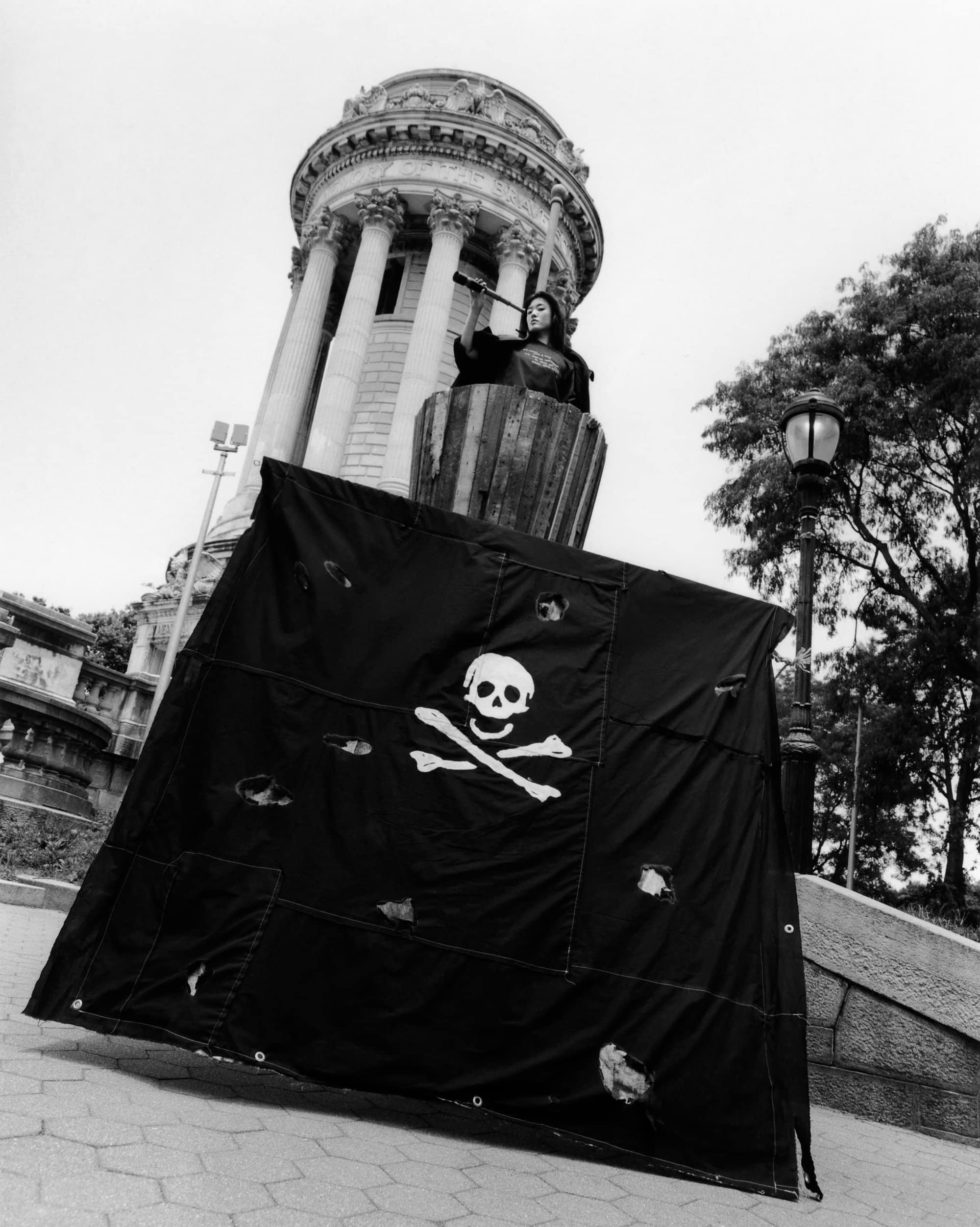 Monochrome image captures a figure standing atop a platform in a barrel, holding a spyglass, with a pirate flag in the foreground.
