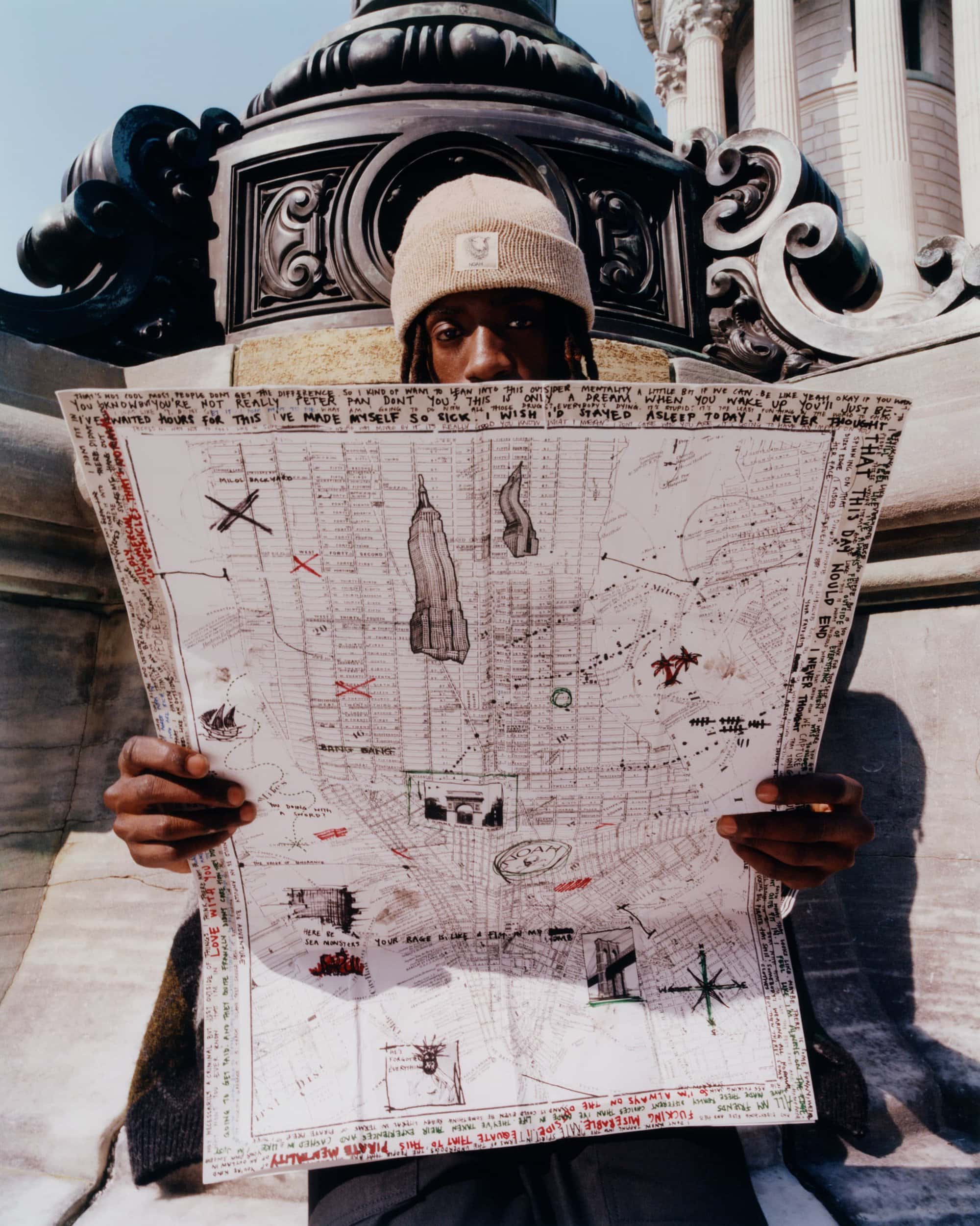 A person with locs and a beanie hat holds a detailed map in front of a building with ornate columns, creating a visual focus on travel.