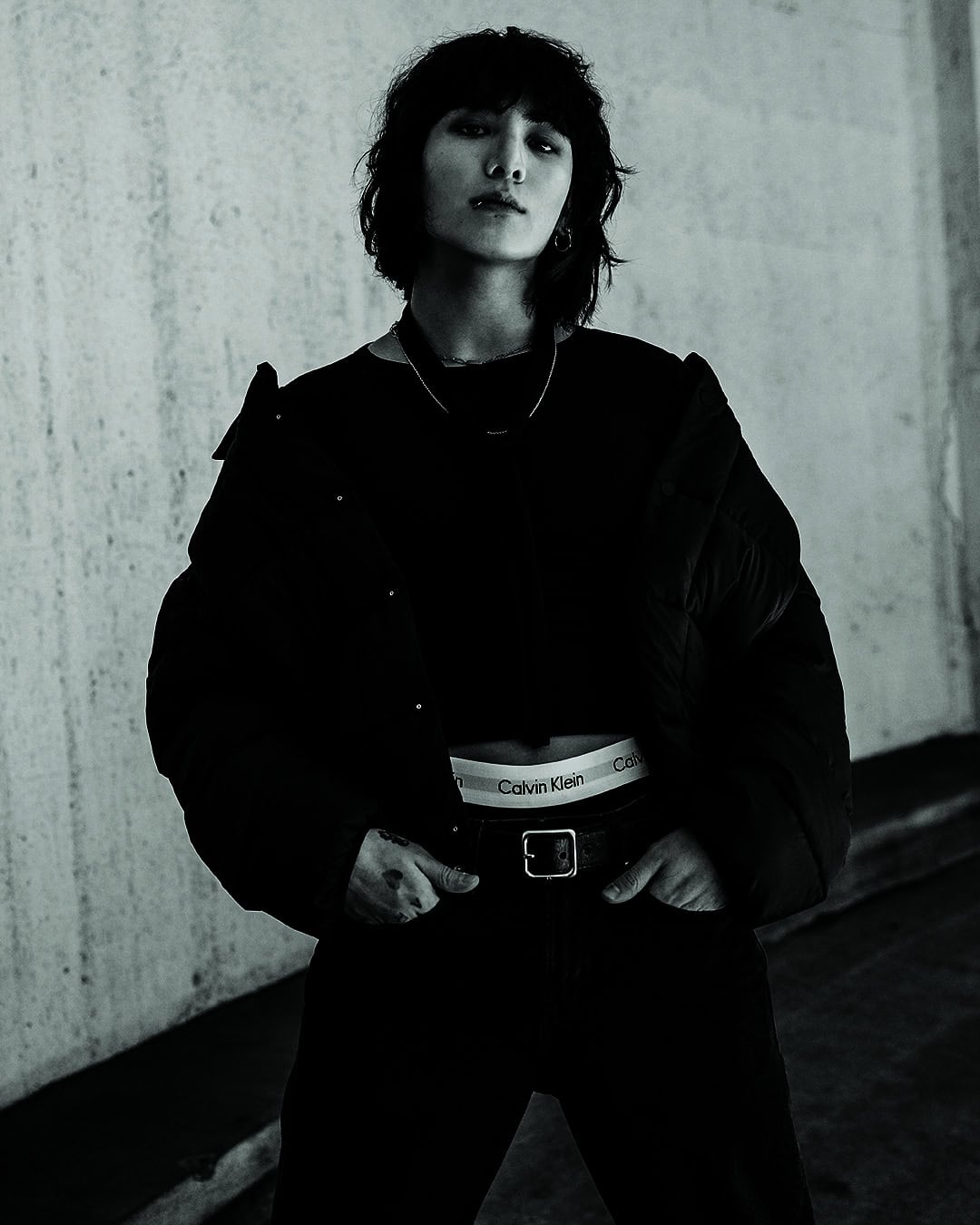 Jung Kook wearing a black puffer jacket and Calvin Klein underwear waistband against a concrete wall.