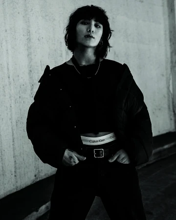 Jung Kook wearing a black puffer jacket and Calvin Klein underwear waistband against a concrete wall.