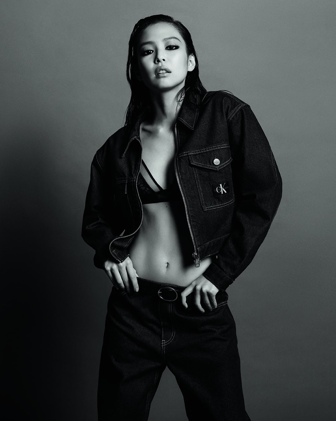 Jennie posing in an unbuttoned black denim jacket and bra for Calvin Klein against a studio backdrop.