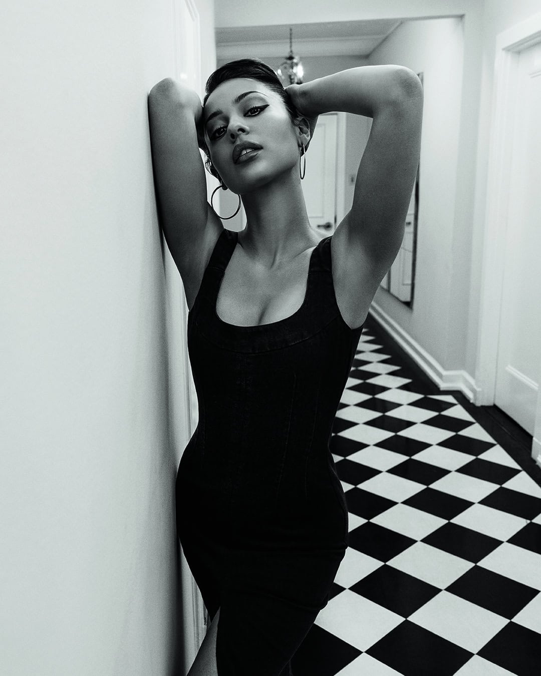 Black and white portrait of a woman in a black dress leaning against a wall, with a checkerboard floor in the background.