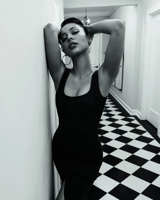 Black and white portrait of a woman in a black dress leaning against a wall, with a checkerboard floor in the background.