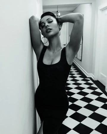 Black and white portrait of a woman in a black dress leaning against a wall, with a checkerboard floor in the background.