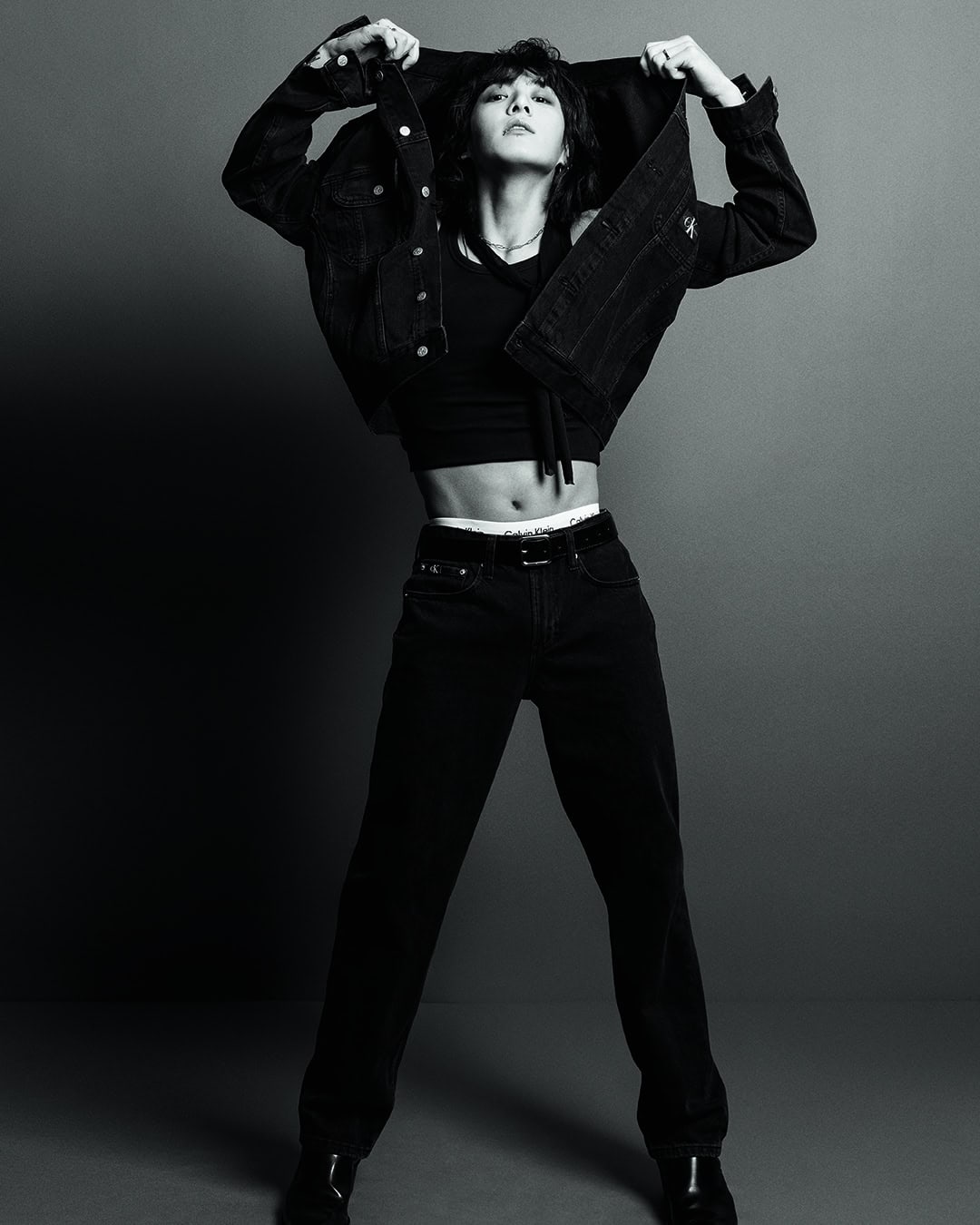 Monochrome portrait of an adult female model in black denim jacket, top, and jeans, striking a confident pose against a grey background.