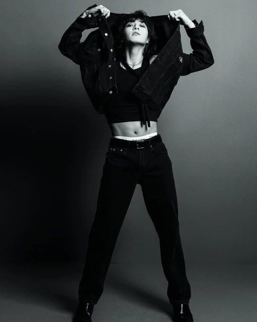 Monochrome portrait of an adult female model in black denim jacket, top, and jeans, striking a confident pose against a grey background.