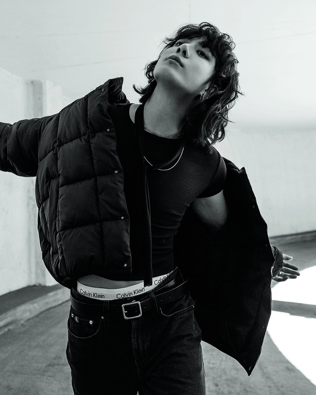 Black and white fashion portrait of a person with dark hair wearing a jacket, black shirt, and jeans in a neutral-tone indoor setting.