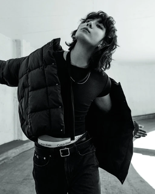 Black and white fashion portrait of a person with dark hair wearing a jacket, black shirt, and jeans in a neutral-tone indoor setting.