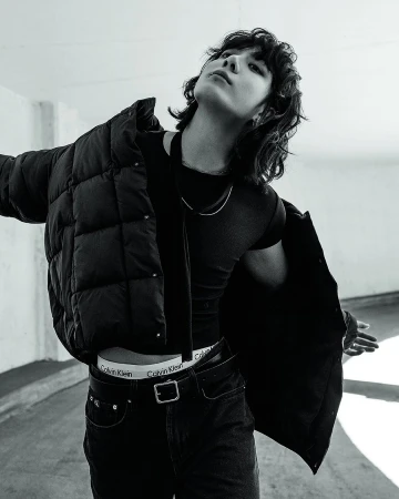 Black and white fashion portrait of a person with dark hair wearing a jacket, black shirt, and jeans in a neutral-tone indoor setting.