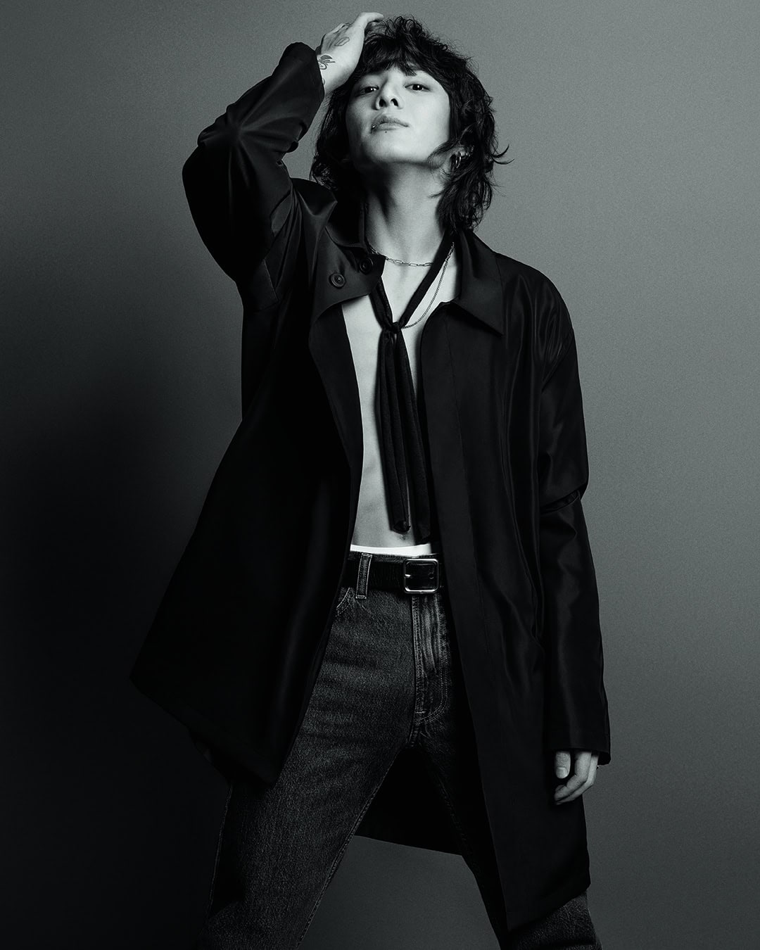Jungkook in a black coat and jeans, posing with one hand on his hair, from the Calvin Klein Fall 2023 campaign.