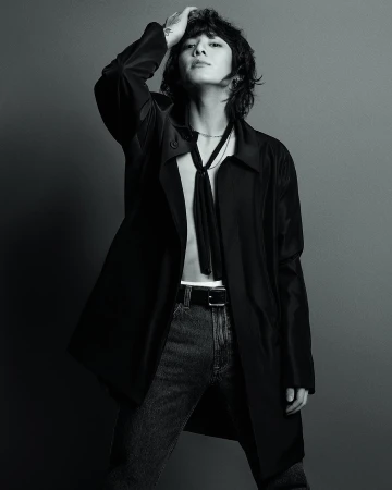 Jungkook in a black coat and jeans, posing with one hand on his hair, from the Calvin Klein Fall 2023 campaign.