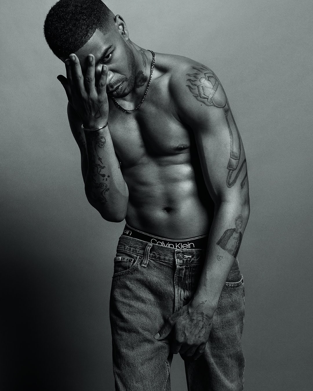 Monochrome portrait of a muscular adult male with tattoos, wearing jeans, and holding his hand to his forehead in a photo studio setting.