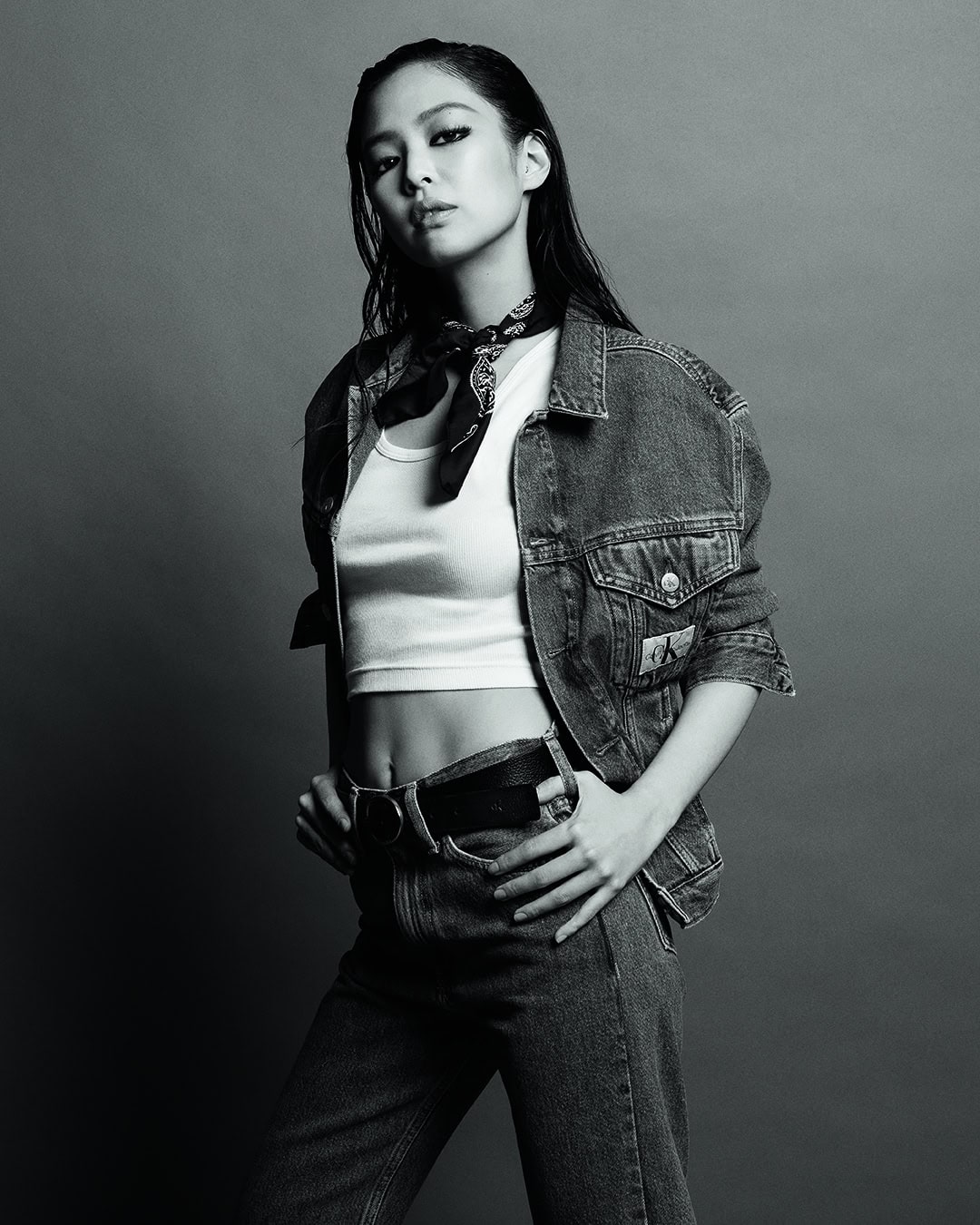Jennie posing in a denim jacket and white tank top against a plain grey studio backdrop for Calvin Klein