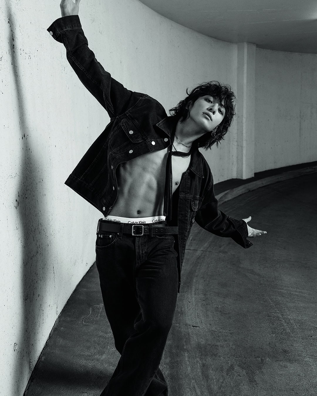 Monochrome portrait of a slender adult male with long, wavy hair and an open denim jacket, posing in an underground setting.