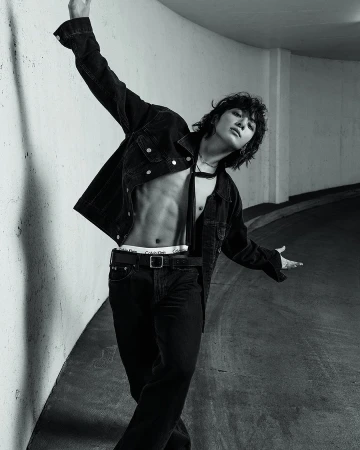 Monochrome portrait of a slender adult male with long, wavy hair and an open denim jacket, posing in an underground setting.