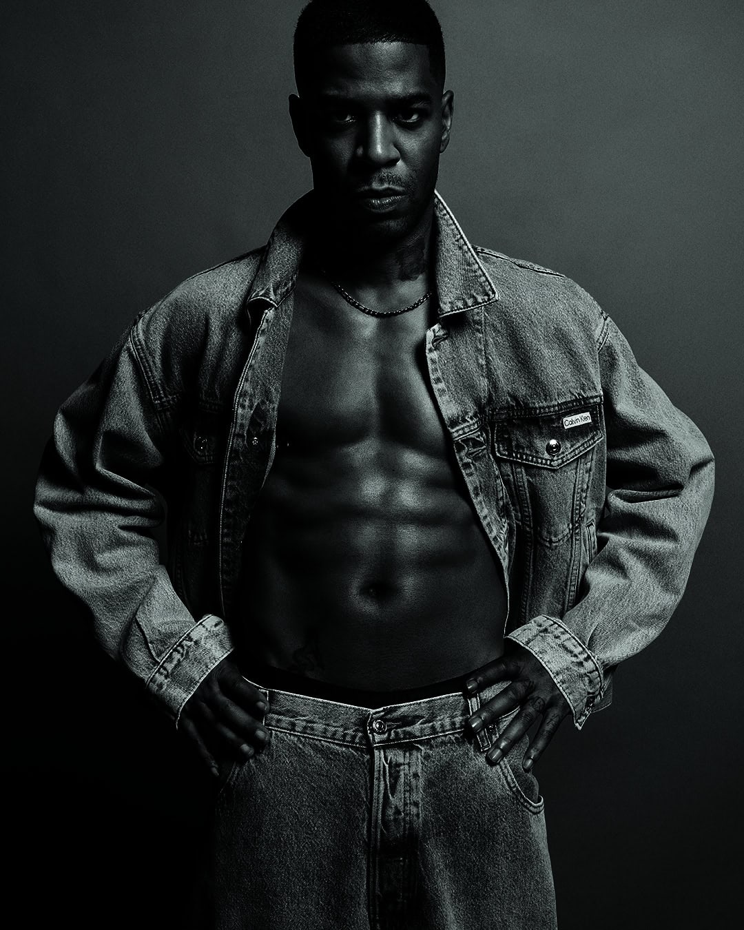 Portrait of muscular African-American male posing in denim jacket and jeans against a grey backdrop in a studio environment.