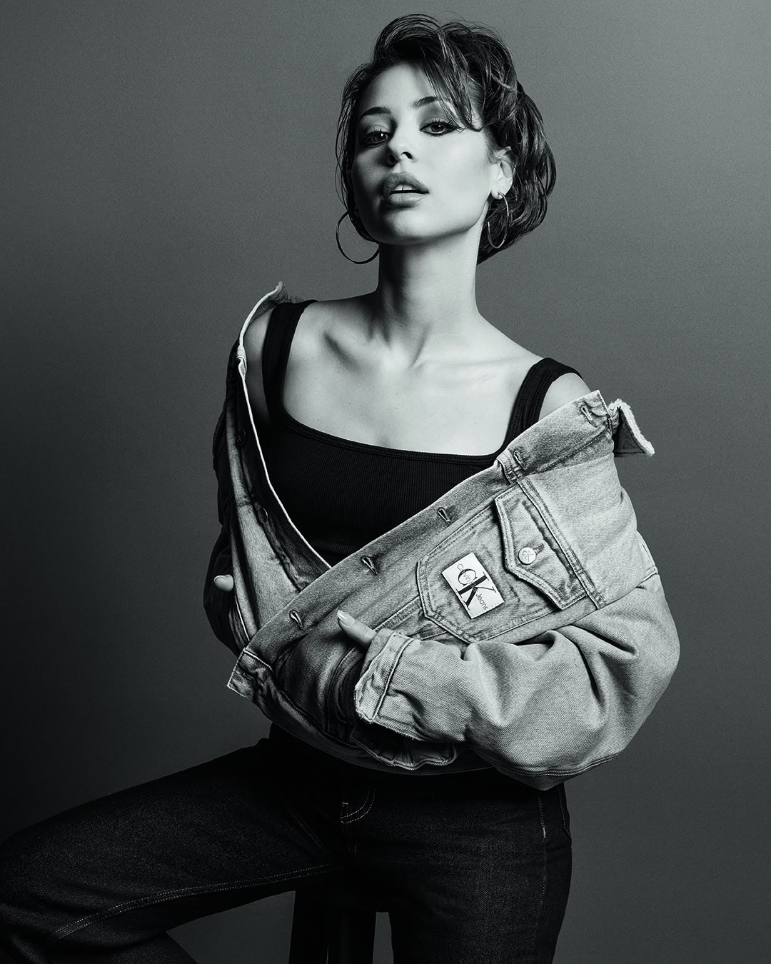 Monochrome portrait of a young woman in a tank top and denim jacket, gazing directly at the camera with subtle makeup and hoop earrings.