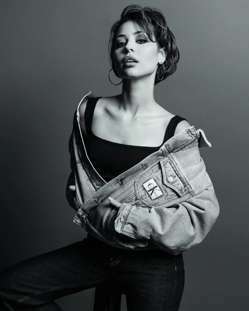 Monochrome portrait of a young woman in a tank top and denim jacket, gazing directly at the camera with subtle makeup and hoop earrings.