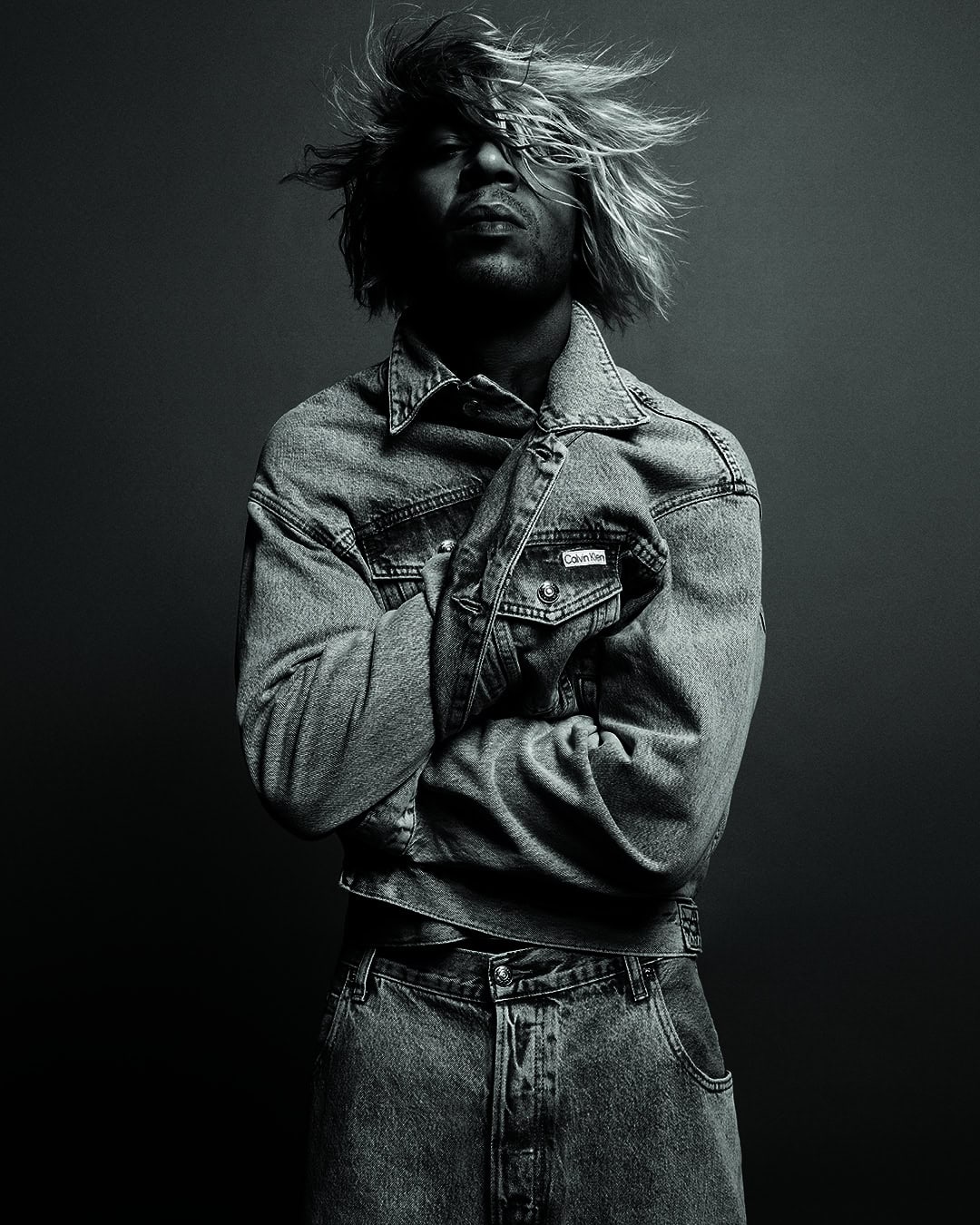Monochrome portrait of an adult male with fair hair, wearing a denim jacket and jeans, arms crossed, against a dark background.