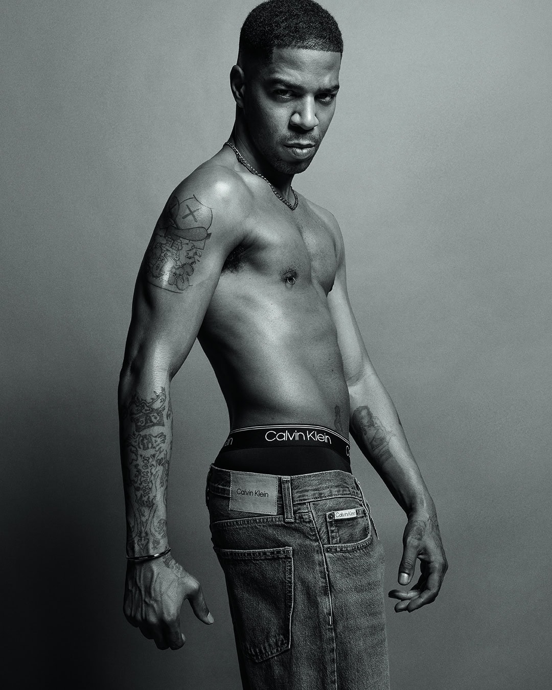 Black and white portrait of muscular adult male with tattoos, wearing jeans and Calvin Klein underwear, in front of grey studio background.