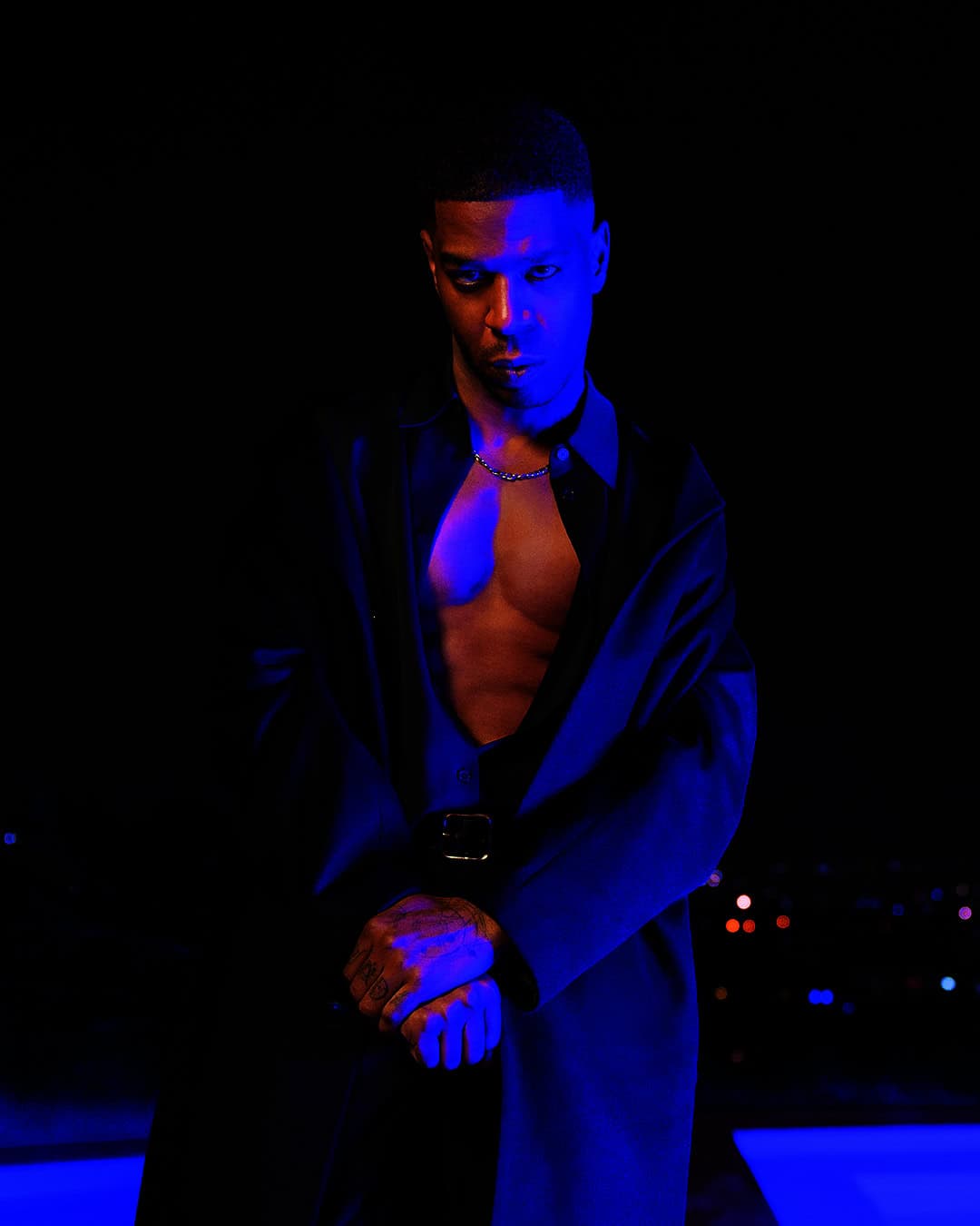 Portrait of a dark-skinned adult male with a short haircut, wearing an open black shirt and necklace, posing indoors with blue lighting.