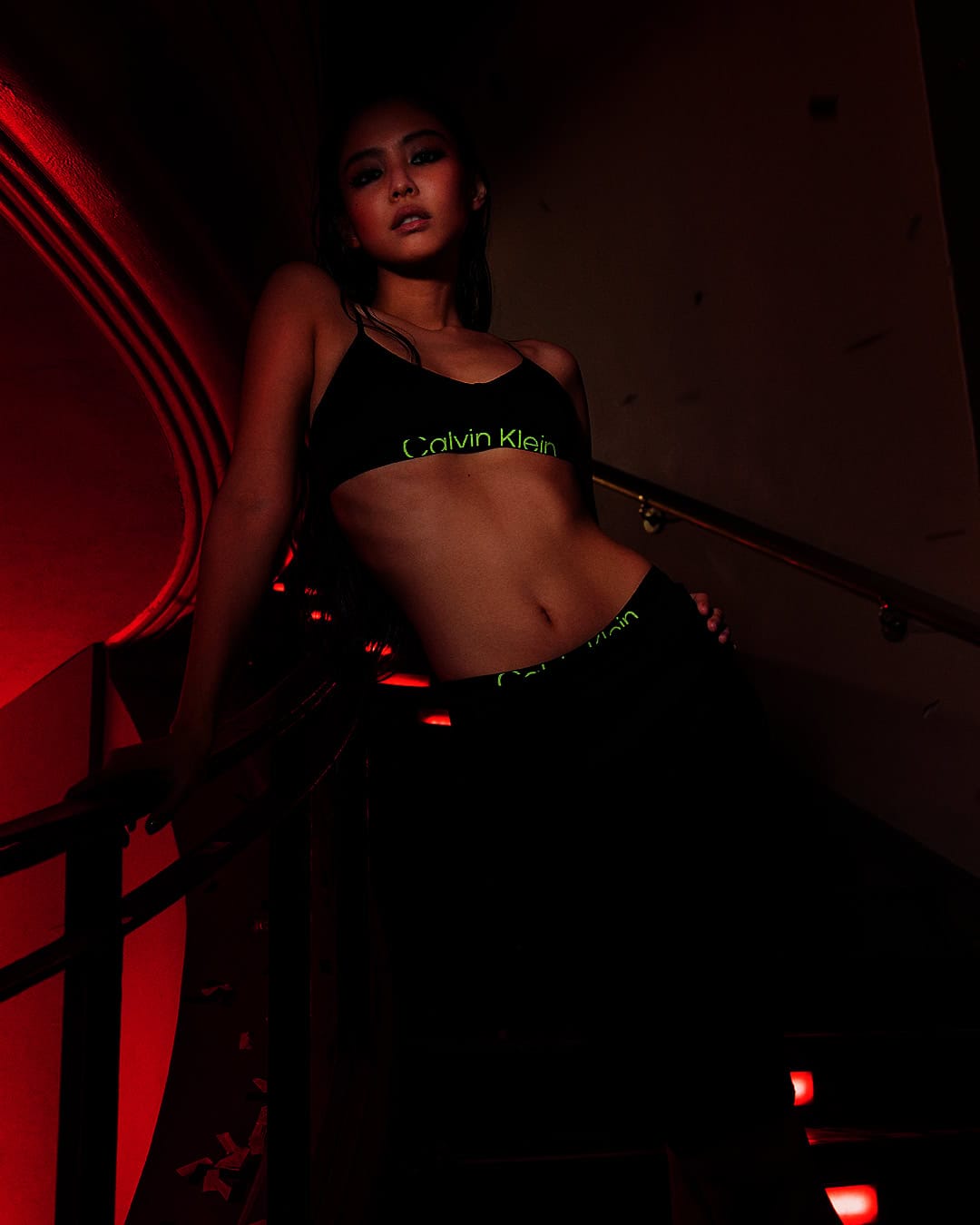 Athletic woman in Calvin Klein underwear and bra poses on red-lit stairs.  Fitness and fashion meet in dim indoor setting.