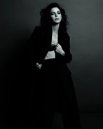 Elegant woman in black blazer and pants poses in a studio setting with dramatic lighting, showcasing a sophisticated fashion style.