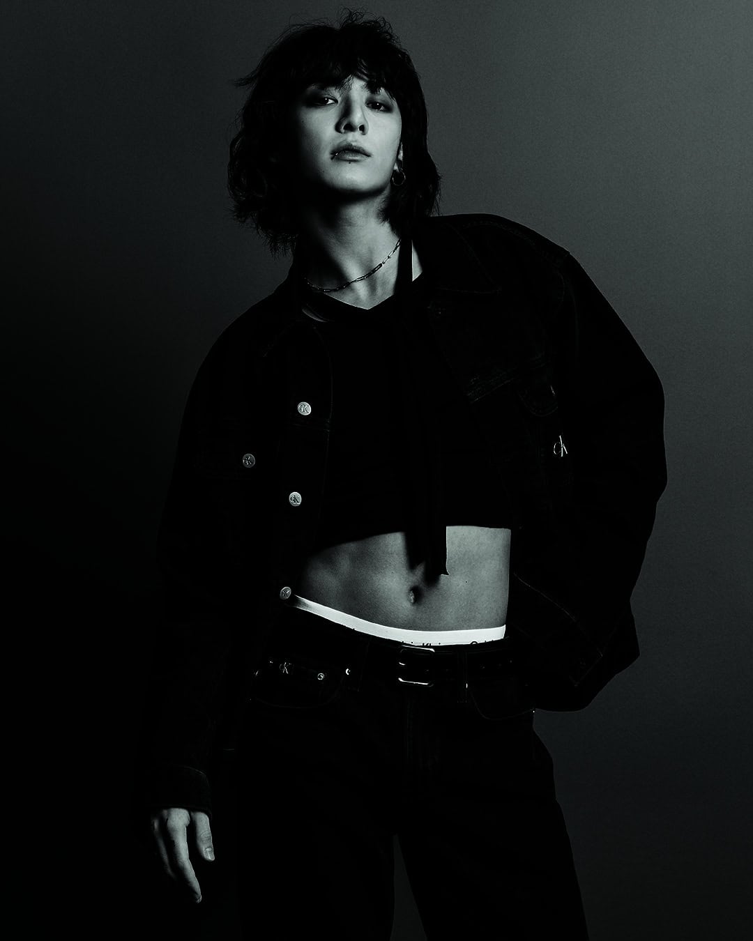 Fashionable person posing indoors with a black crop top and jeans in a photo studio with dark lighting and a gray background.