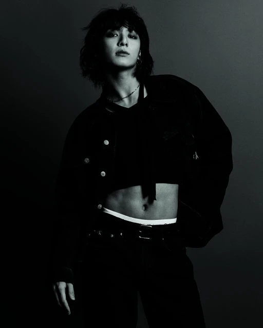 Fashionable person posing indoors with a black crop top and jeans in a photo studio with dark lighting and a gray background.