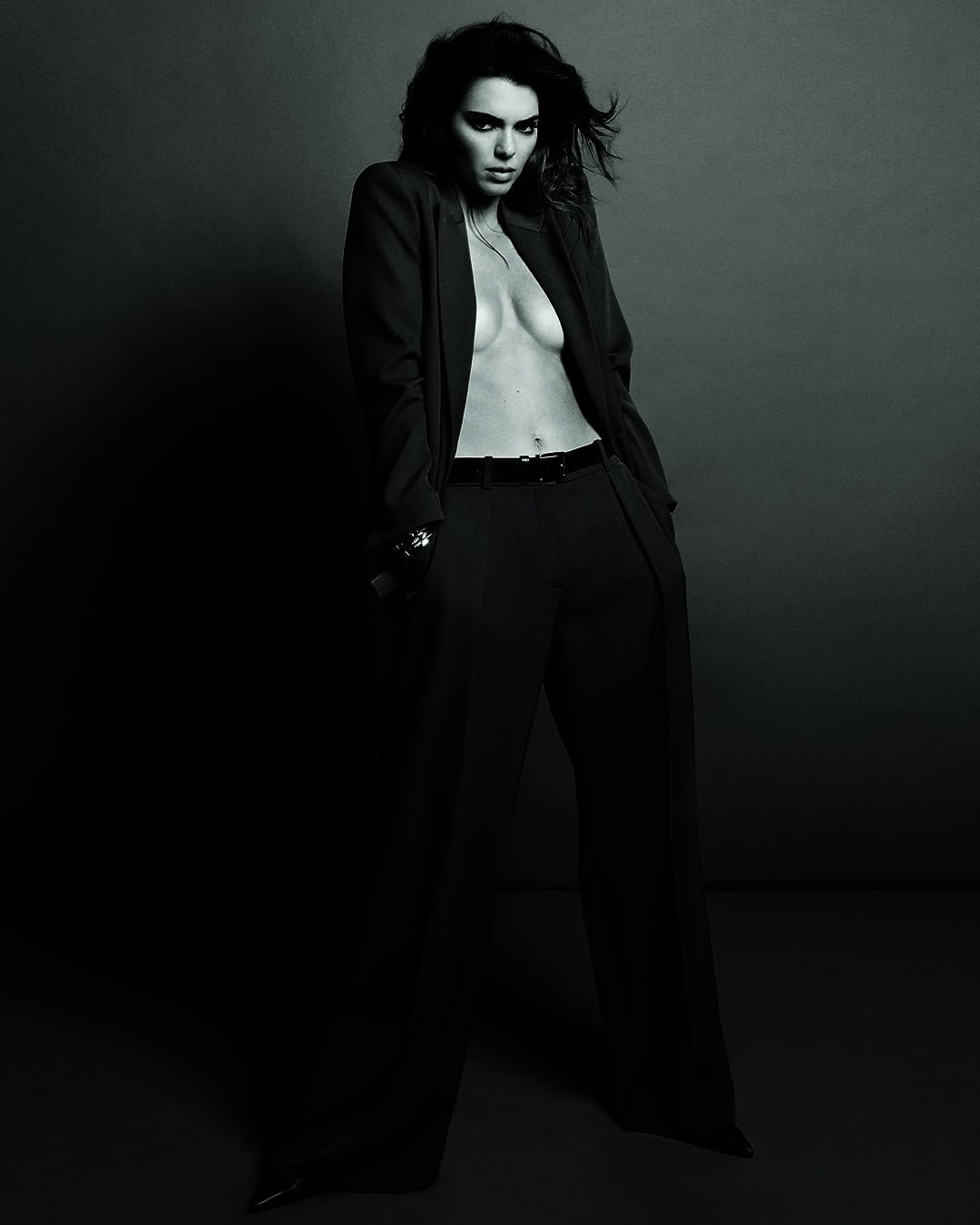 A woman in a black suit stands in a studio, looking at the camera. The image is shot in black and white with a high contrast.