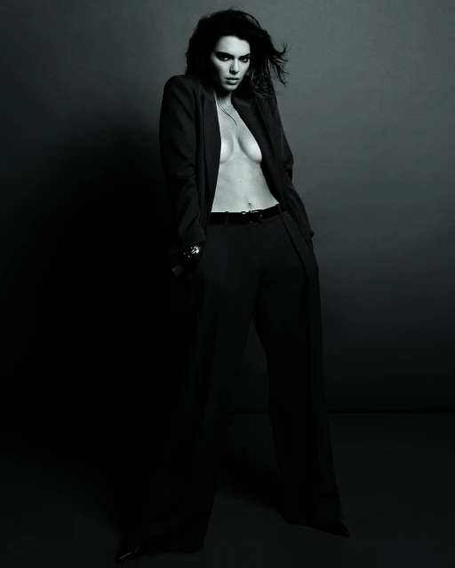 A woman in a black suit stands in a studio, looking at the camera. The image is shot in black and white with a high contrast.
