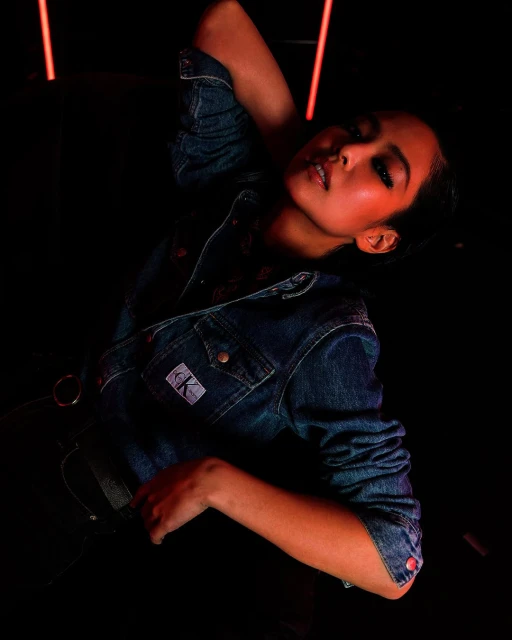 Portrait of a young woman in a denim jacket posing indoors against a dark backdrop with neon light accents for a fashion editorial.