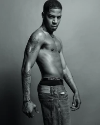 Kid Cudi posing shirtless in Calvin Klein jeans and exposed underwear waistband against a grey studio backdrop.