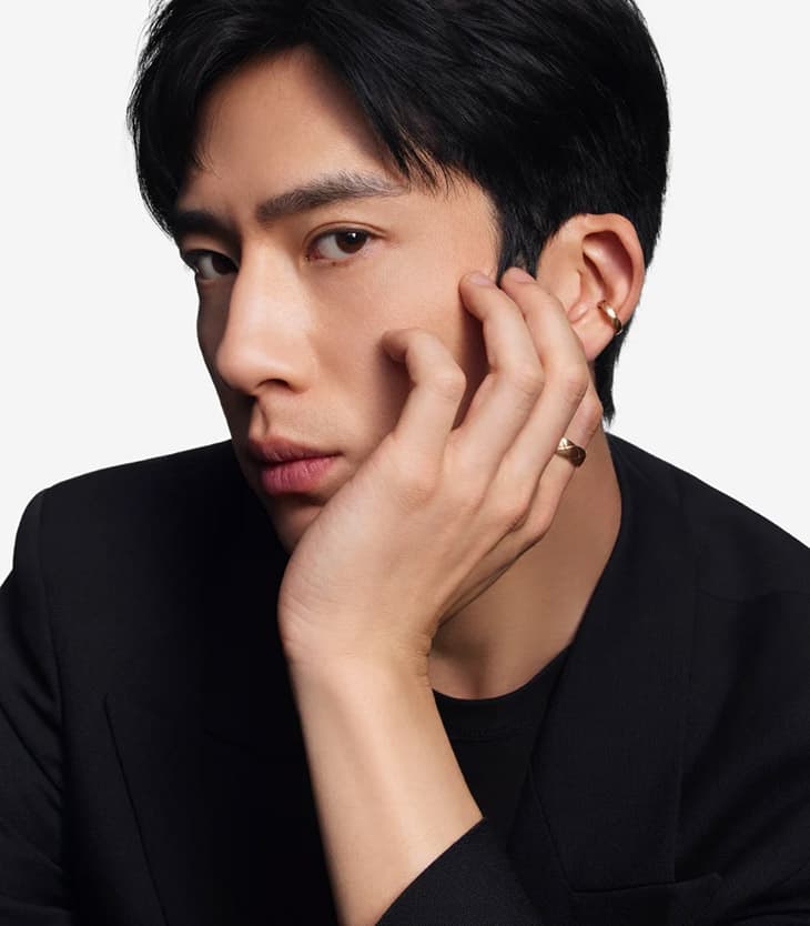 Headshot of a young adult Asian man in a dark blazer against a white background, with minimal jewelry and a serious expression.