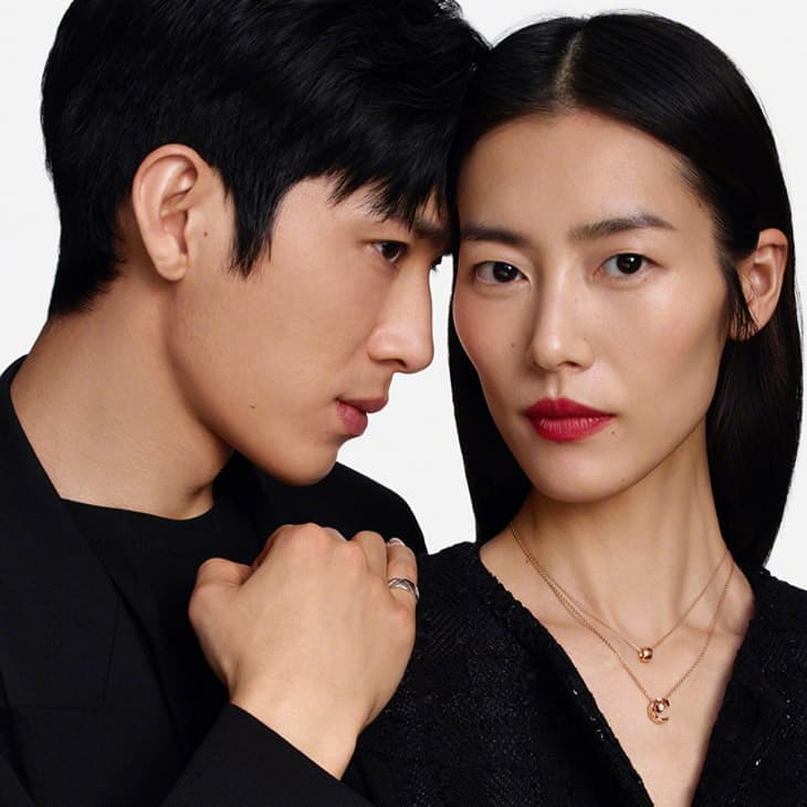Close-up portrait of an Asian couple, a man and a woman, in dark attire against a light background, with focus on their faces and jewelry.