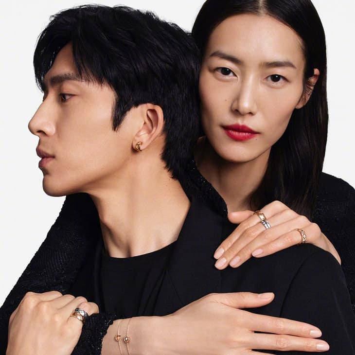 Portrait of a stylish Asian couple wearing jewellery. The woman has her arms around the man, both are in black attire against a white background.