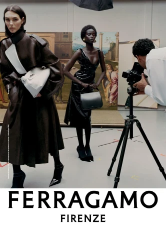Two female fashion models in a studio setting wearing Ferragamo clothing and carrying handbags, photographed by a photographer with a camera.