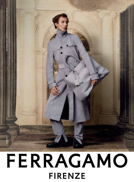 Man in a grey trench coat holds a matching grey tote bag in front of an ornate, classically-inspired painted backdrop in a fashion shot.