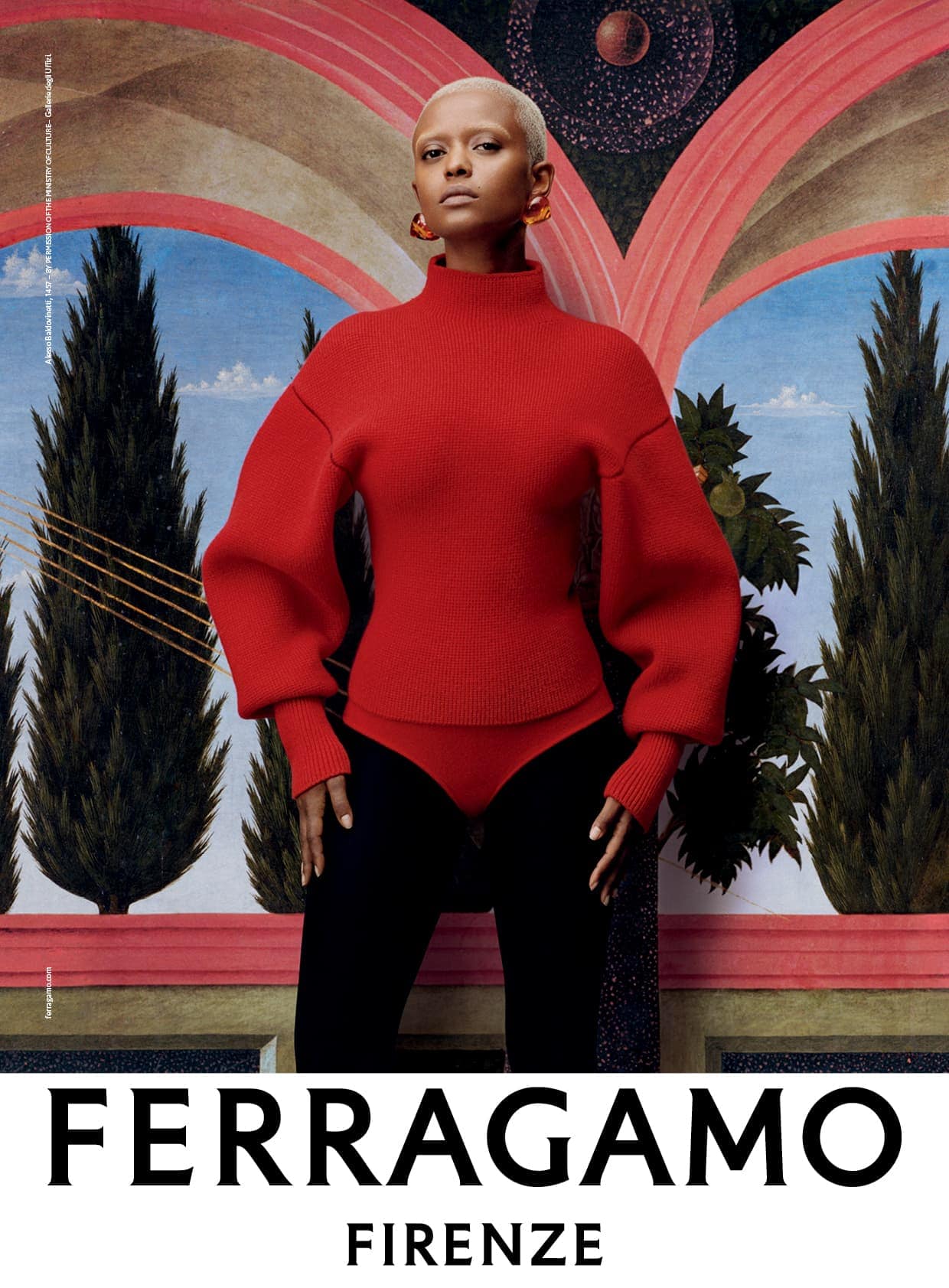 Fashion shot of a woman with blonde hair wearing a red turtleneck sweater and black pants, standing against a scenic backdrop.