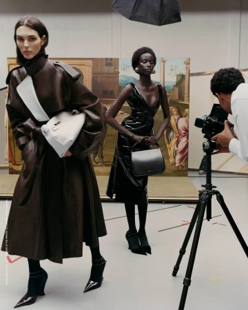 Two fashion models pose with handbags in a photo studio; a photographer captures the scene with vintage art paintings in the backdrop.
