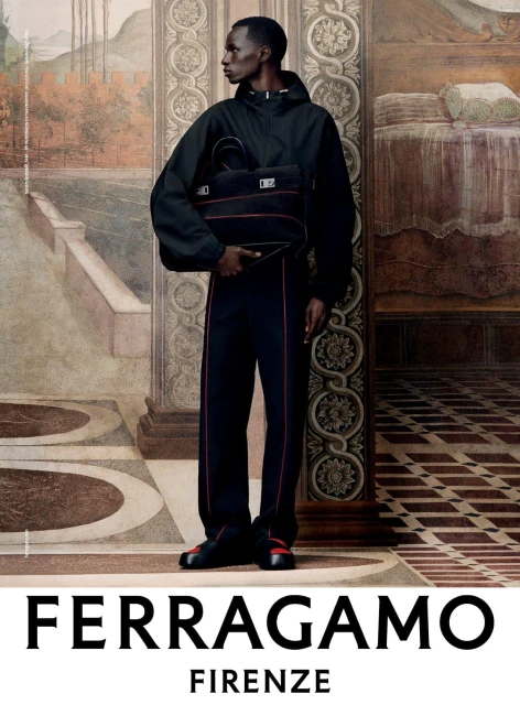 A fashion shot featuring a Black male model in a dark tracksuit holding a bag, posing in front of a painted wall indoors.