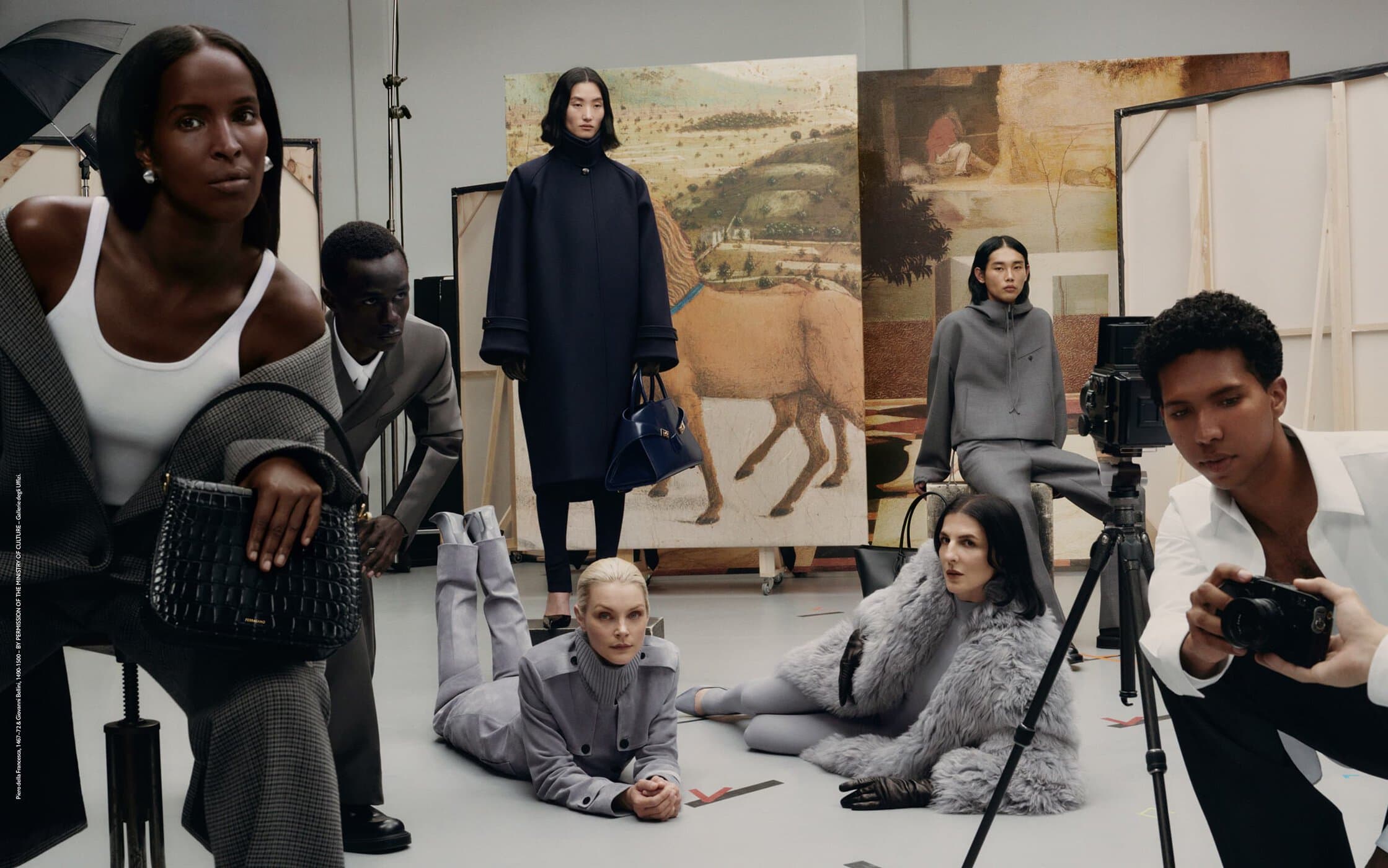 Editorial fashion shot in a photo studio featuring multiple models in modern clothing, handbags, with a photographer and painting backdrop.